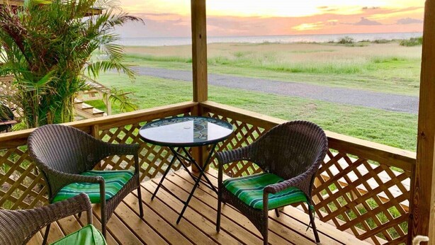 Eine Holzterrasse mit Korbstühlen und einem Tisch, mit Blick auf ein grasbewachsenes Feld und den Ozean bei Sonnenuntergang.