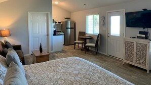 Ein Schlafzimmer mit Bett, Deckenventilator, Nachttisch, Lampe, Kommode, Fernseher und einer Küchenzeile mit Kühlschrank und Fenster.