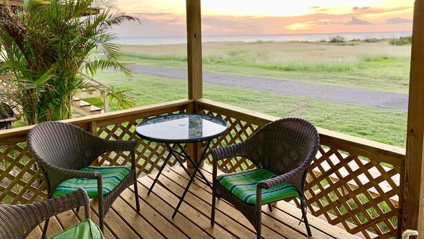 Eine Holzterrasse mit Korbstühlen und einem Tisch, mit Blick auf ein Grasfeld und einen Strand bei Sonnenuntergang.