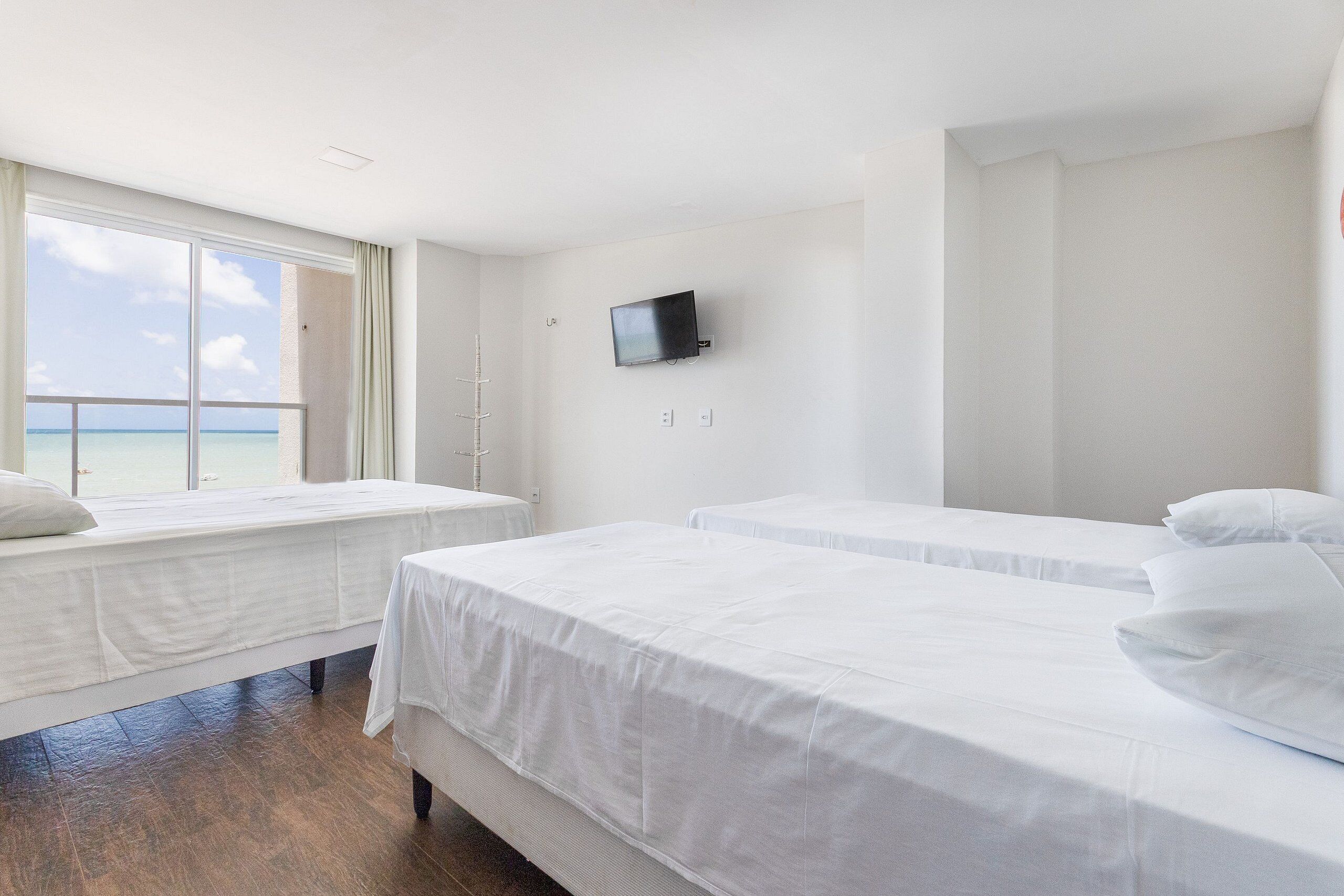A hotel room with two beds, a TV on the wall, and a window with a view of the sea.