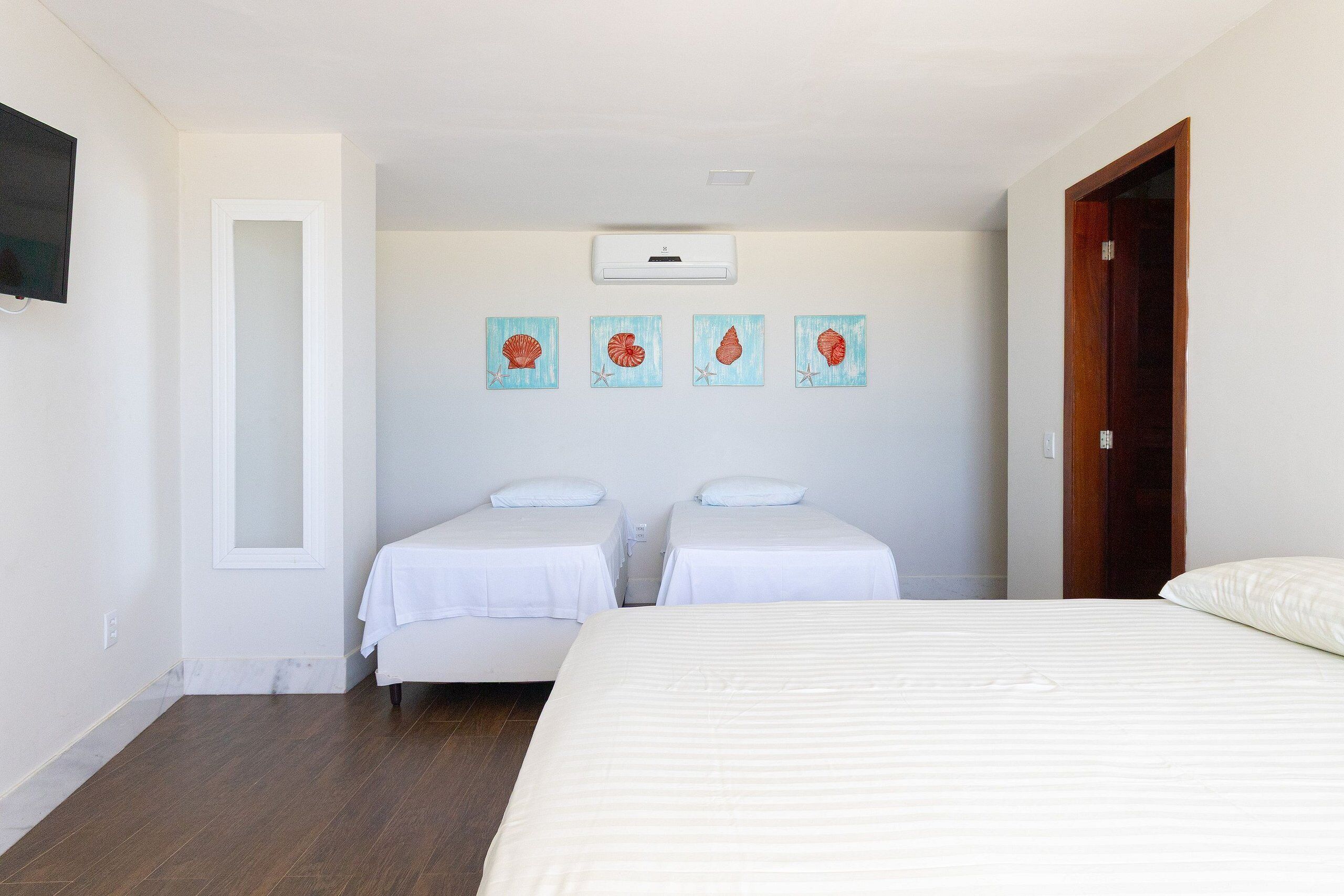 A hotel room with two single beds, a large bed, a flat-screen TV mounted on the wall, and four framed artwork pieces above the beds.