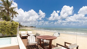 A wooden deck with white lounge chairs and a round table overlooking a beach with clear skies and a pool. - Casa luxuosa a beira mar de Jacumã RN (Ceara-Mirim)