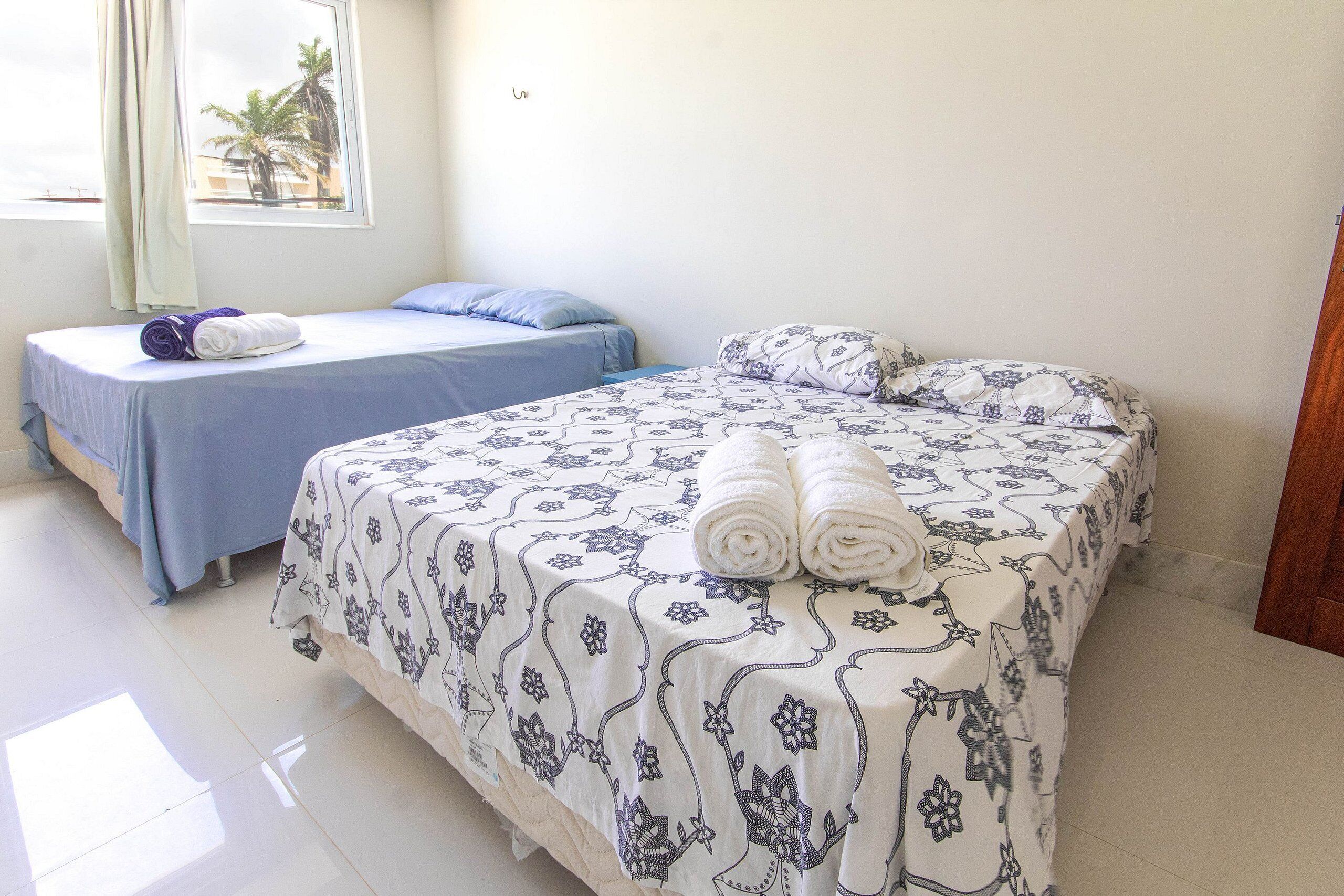 Habitación de hotel con dos camas, una con cubrecama azul y otra con cubrecama blanca con motivos florales azules. Hay toallas sobre las camas y una ventana con cortinas.