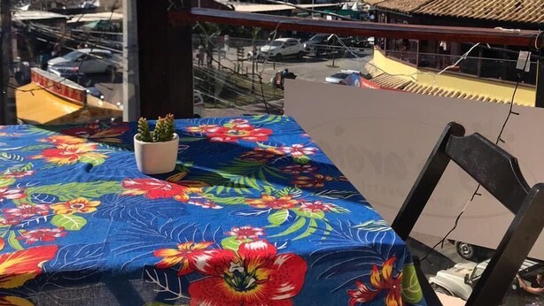 A table with a floral tablecloth, a potted plant, and a view of a beach and marina. - Pe Na Areia Beach Hostel (Araruama)