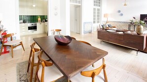 Un vaste espace de vie comprenant une table à manger en bois, des chaises et un canapé. On aperçoit, au fond, une cuisine avec des meubles blancs et un plan de travail vert.