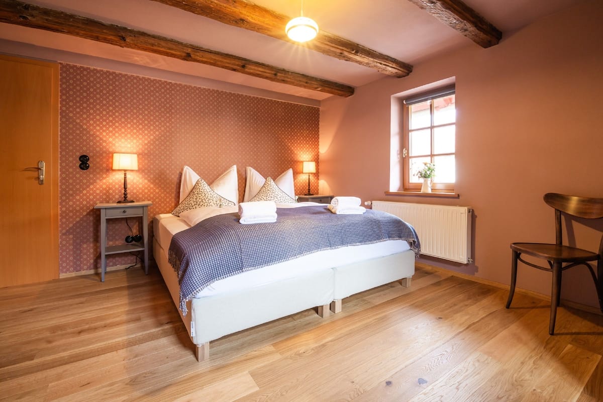 Ein Schlafzimmer mit Holzboden, einem Bett mit gemustertem Überwurf, zwei Nachttischlampen, einem Fenster mit Pflanze und einem Holzstuhl.
