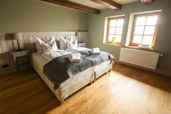 A bedroom with a large bed, wooden floor, a radiator, two windows, and a nightstand with a lamp. - Restaurant & Hotel Frankenberger (Amt Wachsenburg)