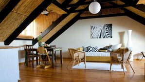 A cozy loft with a high ceiling, exposed wooden beams, a dining area with a table and chairs, a sofa set, and a large abstract painting on the wall. - Sundowner Loft (Mossel Bay)
