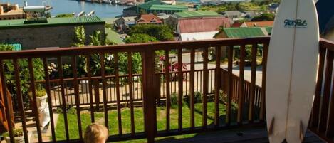 A wooden deck with a child and an adult sitting on a striped lounge chair, overlooking a marina and distant mountains.