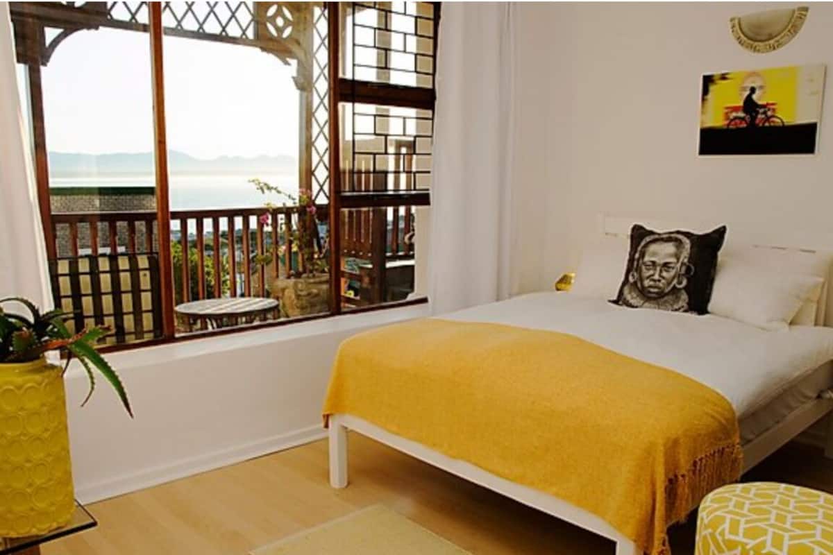 A bedroom with a bed, a window with a view, a yellow plant, and a painting on the wall.