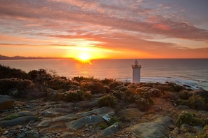 A coastal landscape with a lighthouse, rocky terrain, and a sunset over the ocean. - Sundowner Loft (Mossel Bay)