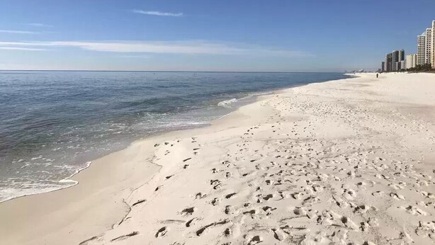 Ein sandiger Strand mit Fußspuren, die zum Wasser führen, ein klarer Himmel und Gebäude im Hintergrund.