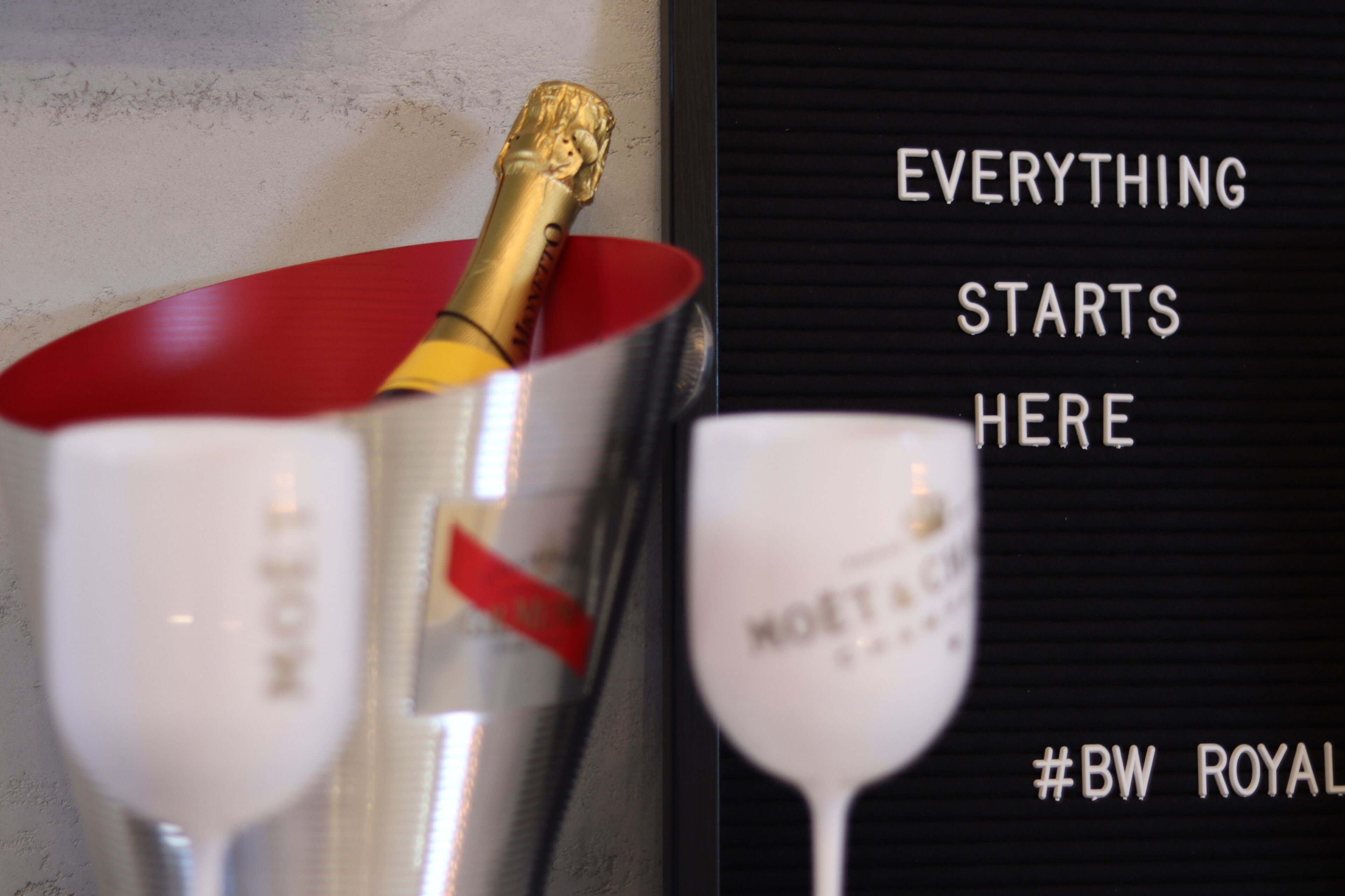 A champagne glass and a bottle with a red foil, and a sign with the text "EVERYTHING STARTS HERE" and "#BW ROYAL".