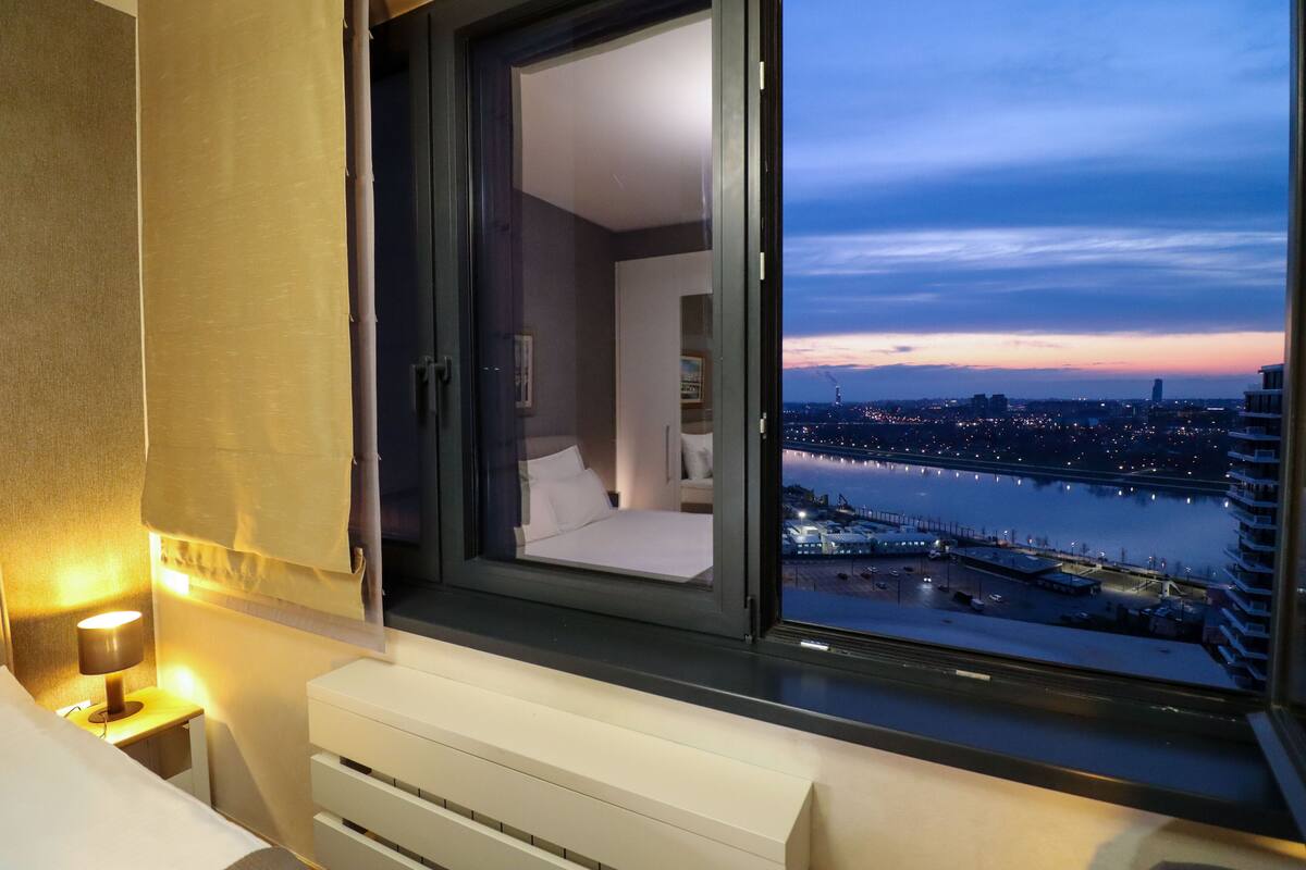 A bedroom with a view of a cityscape and a river at dusk.