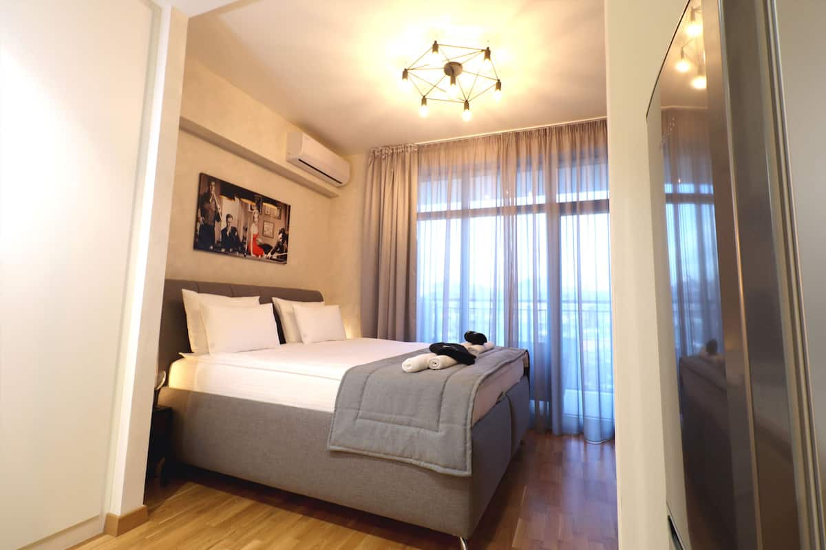 A modern hotel room with a large bed, a ceiling light fixture, a wall-mounted air conditioner, and a window with curtains.