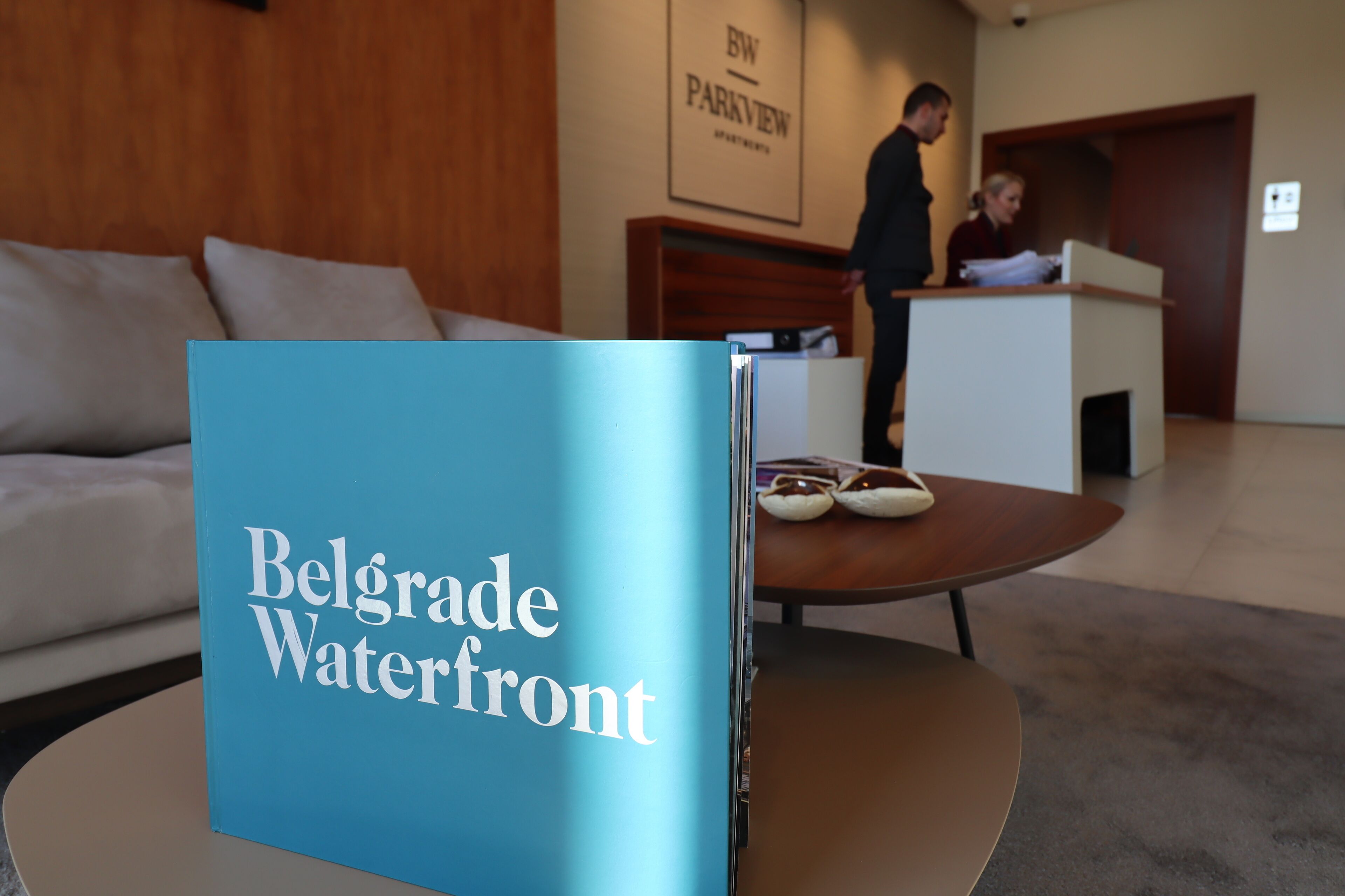A brochure titled "Belgrade Waterfront" in a hotel lobby with a wooden reception desk, a sofa, and a coffee table.