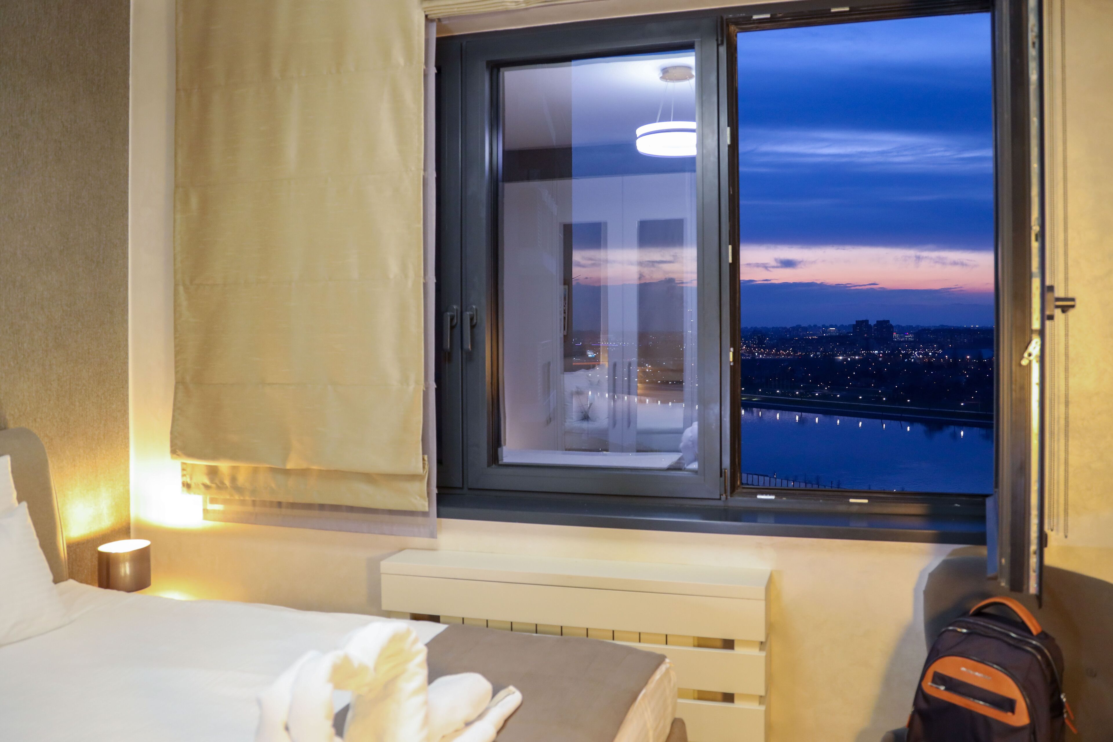 Una camera d'albergo con una grande finestra, un letto, un comodino e una valigia.