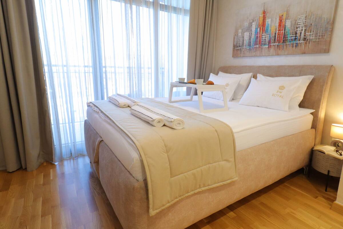 A neatly made bed with a white comforter, two pillows, and a rolled towel. There is a small table with a cup and orange fruit on it. The room has wooden flooring and a large window with sheer curtains.
