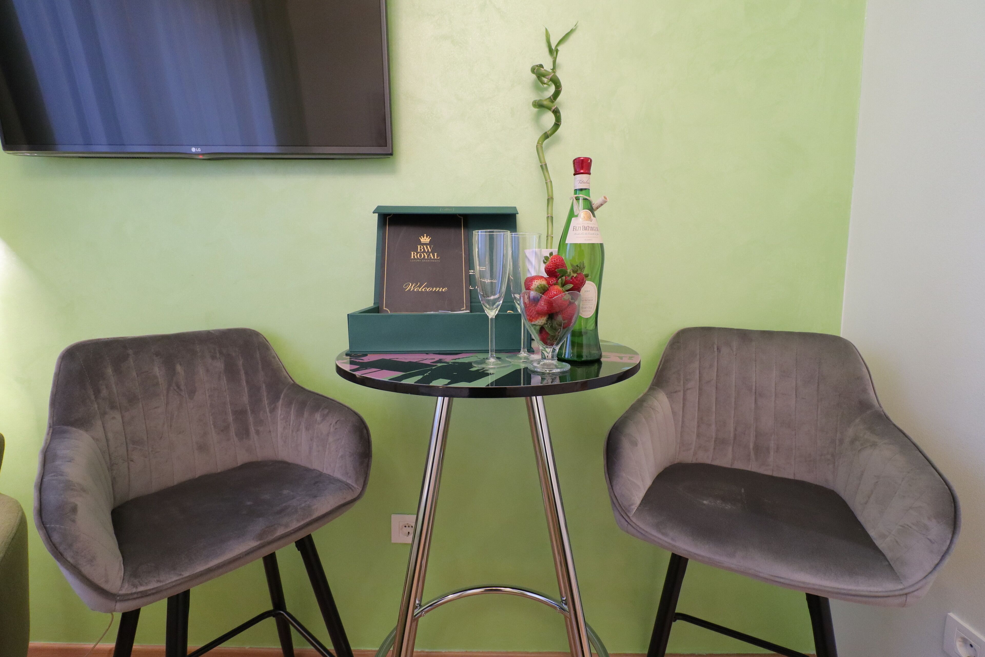 Ein kleiner Tisch mit einem Glas und einer Flasche, zwei graue Stühle und ein gerahmtes Willkommen-Schild.