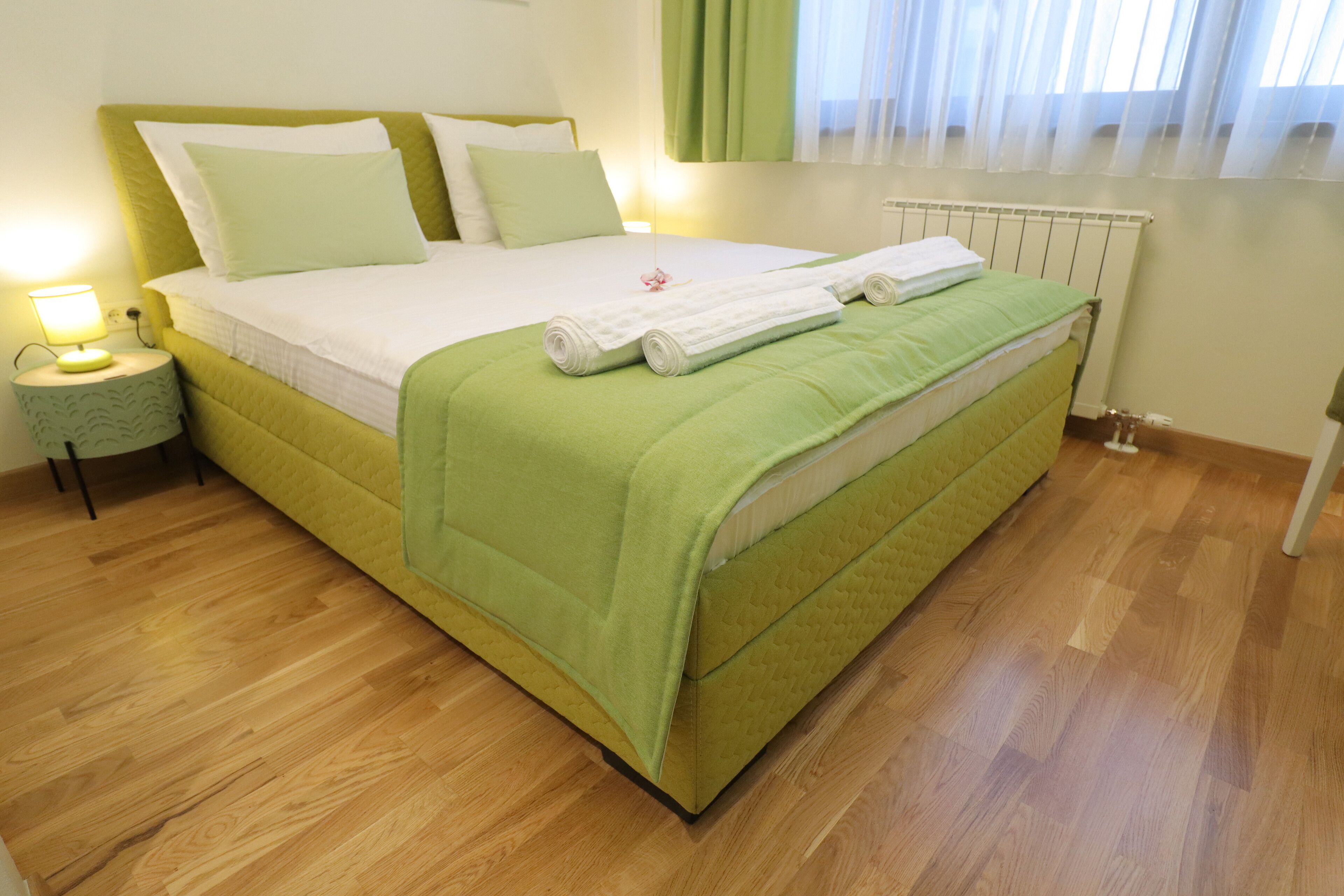 Een net opgemaakt bed met een groene sprei en kussens, een bedlampje en een houten vloer.