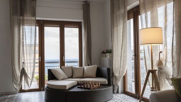 Ett rum med en böjd soffa, ett schackbräde, en lampa och stora fönster med gardiner.