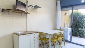 Ein Zimmer mit einem weißen Schreibtisch, gelben Stühlen, einem Fernseher auf einem Regal, einer Waschmaschine und einer Glastür zum Garten.