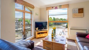 Un salón con televisor, muebles de madera y amplios ventanales con vistas a un río.
