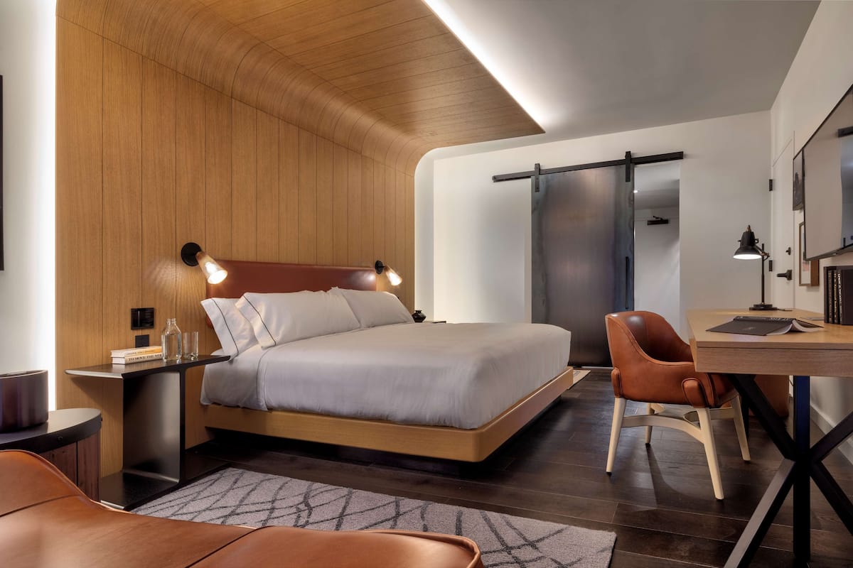 A modern bedroom with a wooden ceiling, a bed with a headboard, bedside lamps, a desk with a chair, and a sliding door.