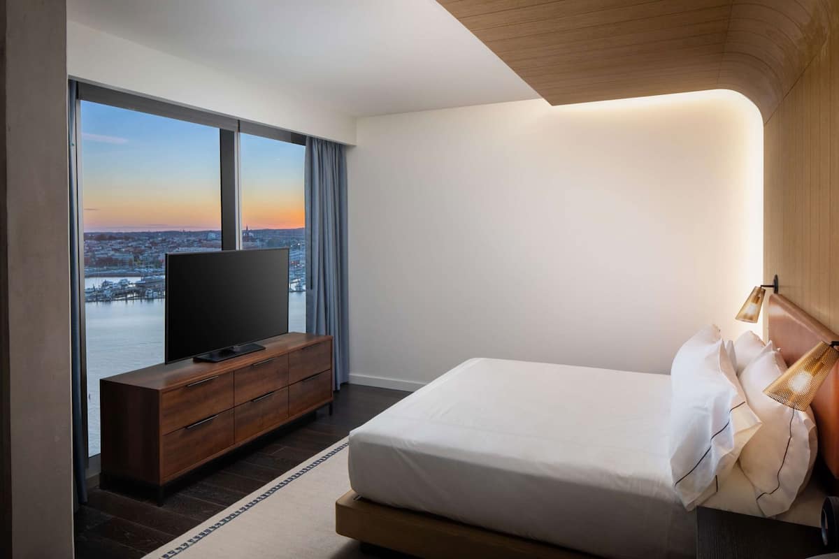 A modern hotel room with a large window, a flat-screen TV on a wooden dresser, a bed with white linens, and a wall-mounted lamp.