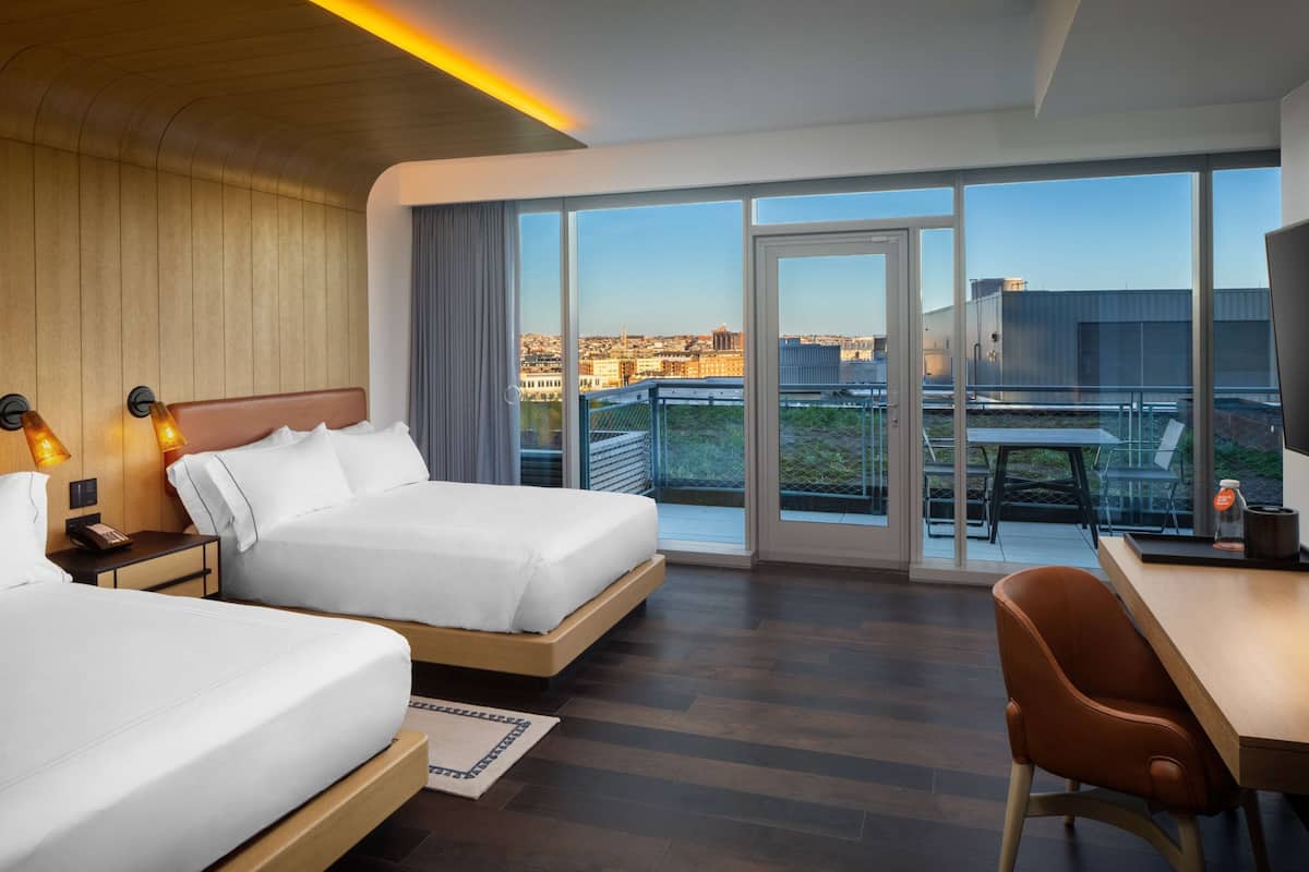 A hotel room with two beds, a desk, and a balcony with a city view.