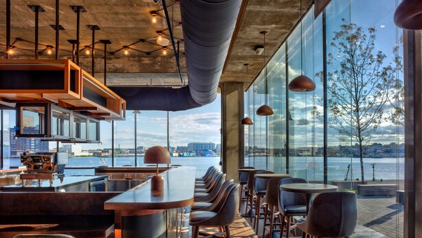 A modern bar area with a view of the waterfront, featuring bar stools, a bar counter, and pendant lighting. - Canopy by Hilton Baltimore Harbor Point (Baltimore)