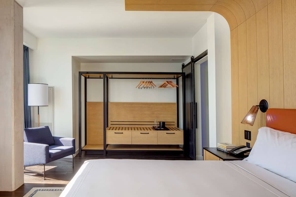 A modern hotel room with a large bed, a desk with a lamp, a chair, and a wardrobe with hanging clothes.