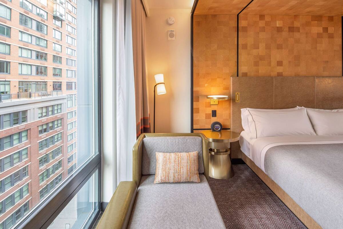 A hotel room with a large window, a bed, a sofa, a bedside table, and a wall-mounted light.