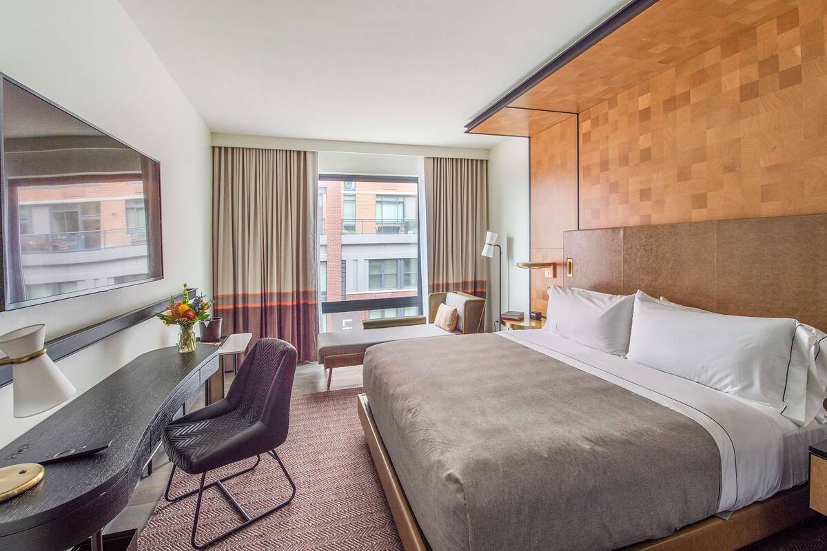 A hotel room with a large bed, a desk, a chair, a lamp, and a view of the city through the window.