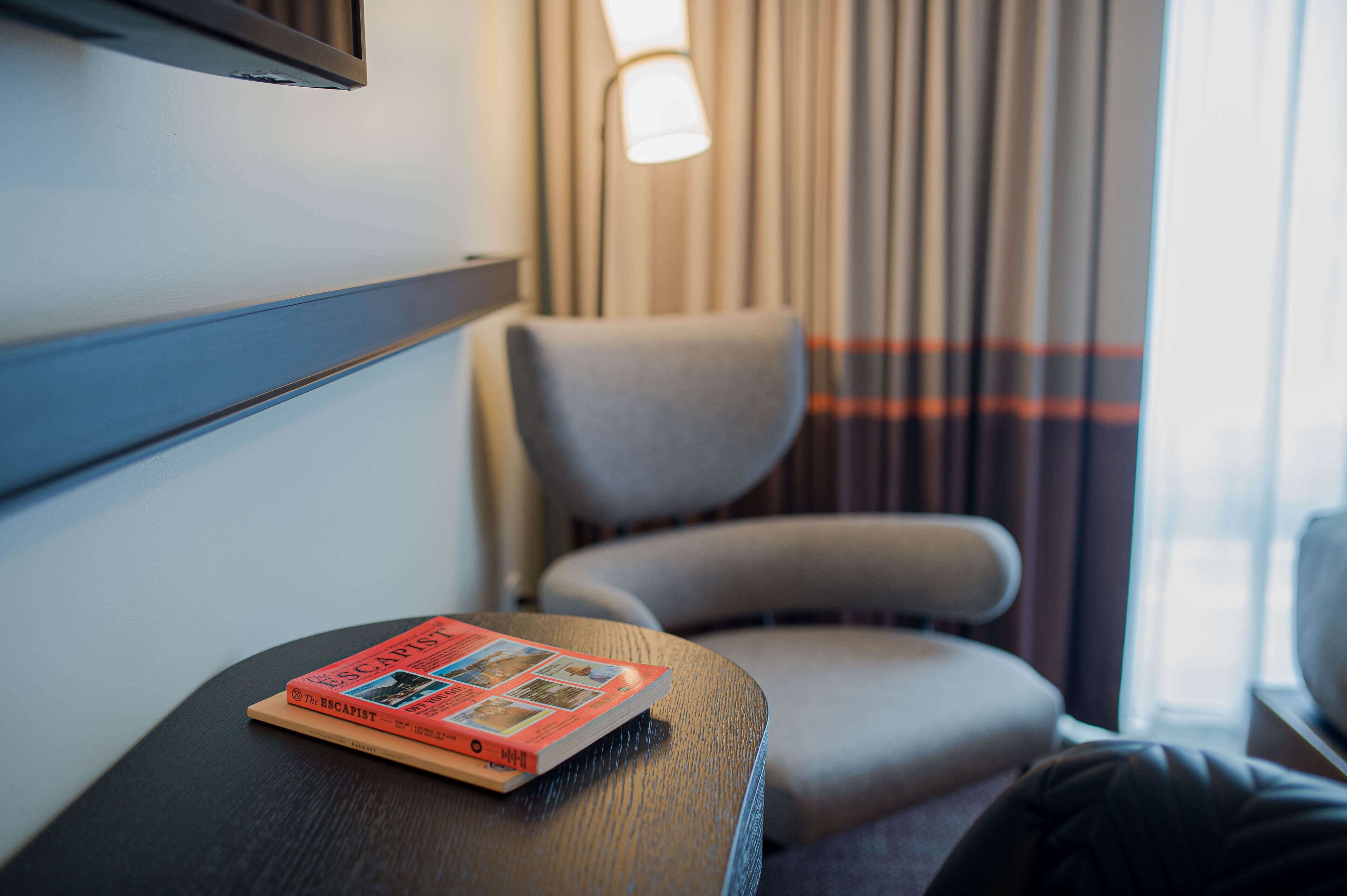 A hotel room with a single armchair, a small table with a book, and a lamp.
