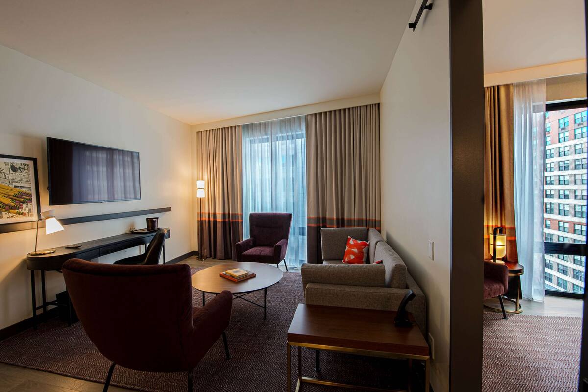 A hotel room with a desk, a flat-screen TV, a sofa, a chair, and a window with curtains.