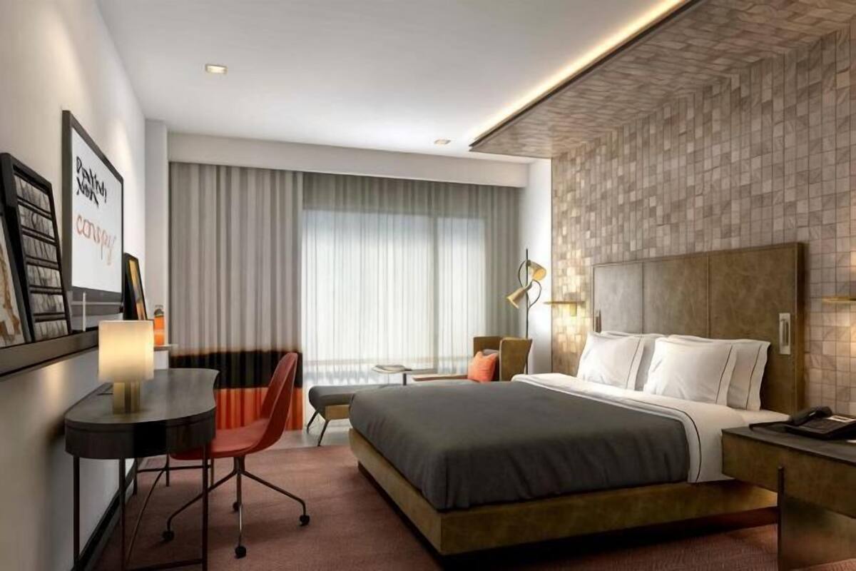 A modern hotel room with a large bed, a desk with a chair, a small table, and a lamp.