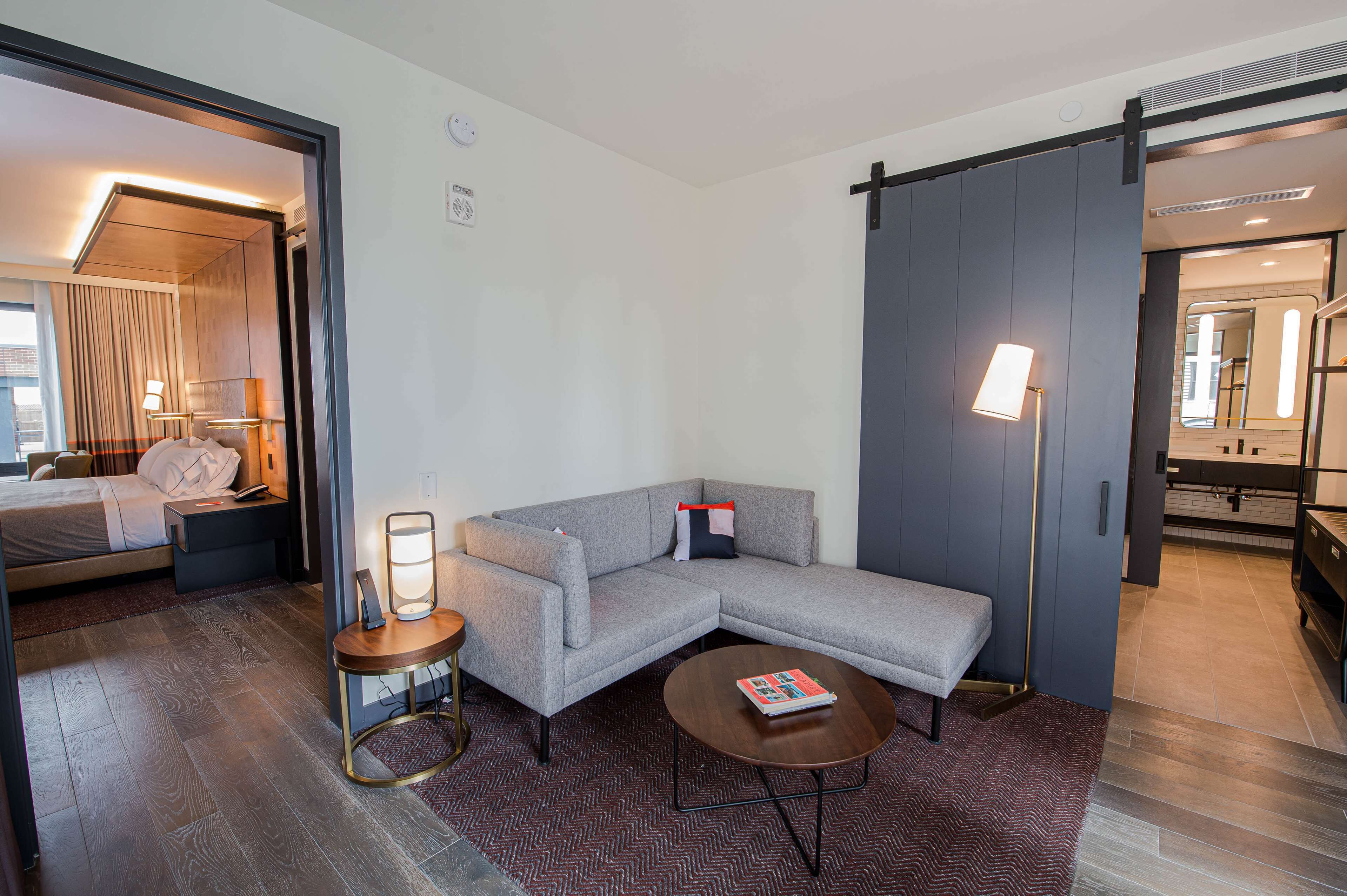 A modern hotel room with a large gray sectional sofa, a small round table with a book, a bedside table with a lamp, and a view of the bathroom through a sliding door.