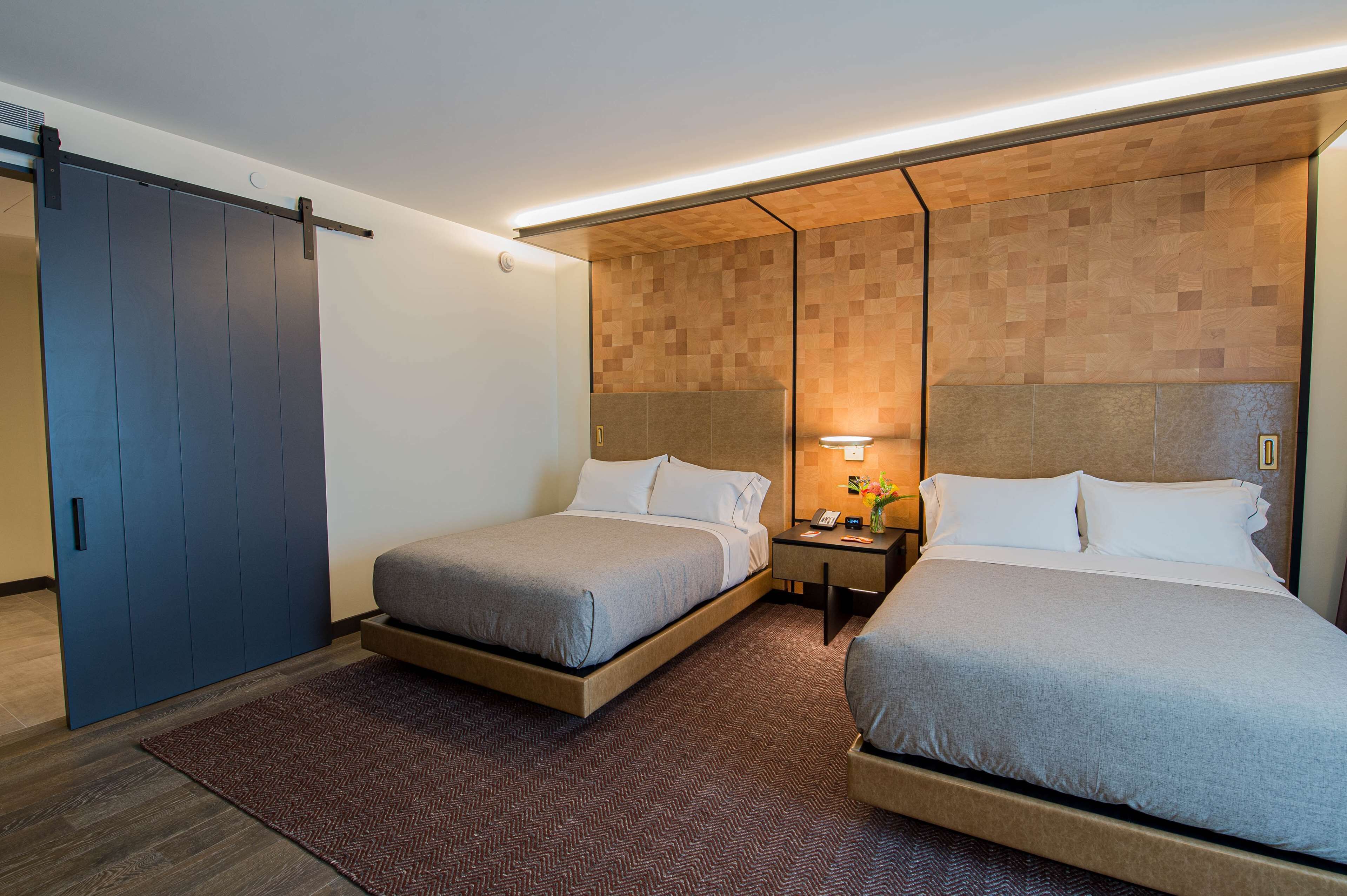 Habitación de hotel con dos camas, cabecero de madera, mesita de noche con lámpara y puerta corredera.