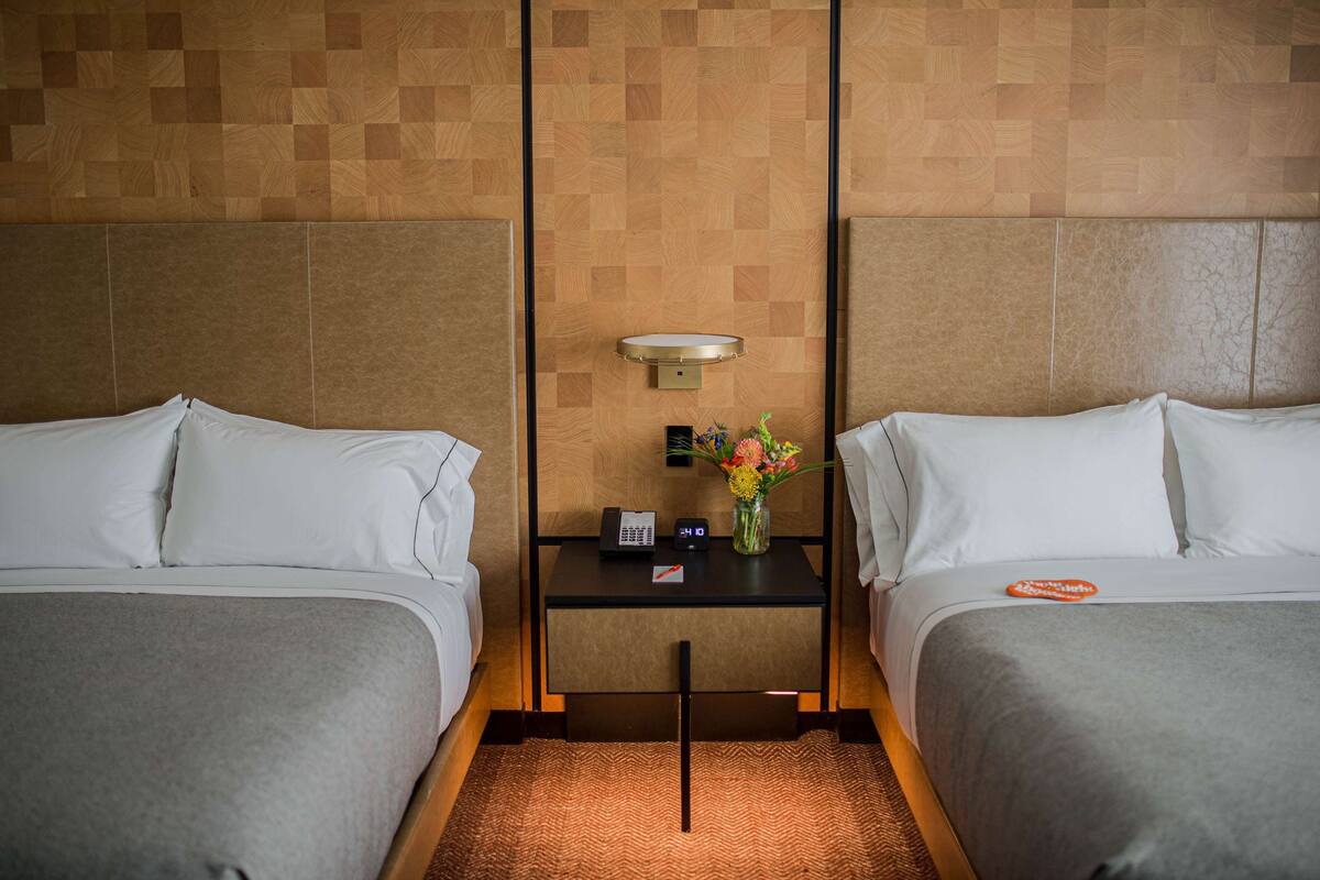 A hotel room with two beds, a nightstand with a phone and a vase of flowers, and a wall-mounted light fixture.
