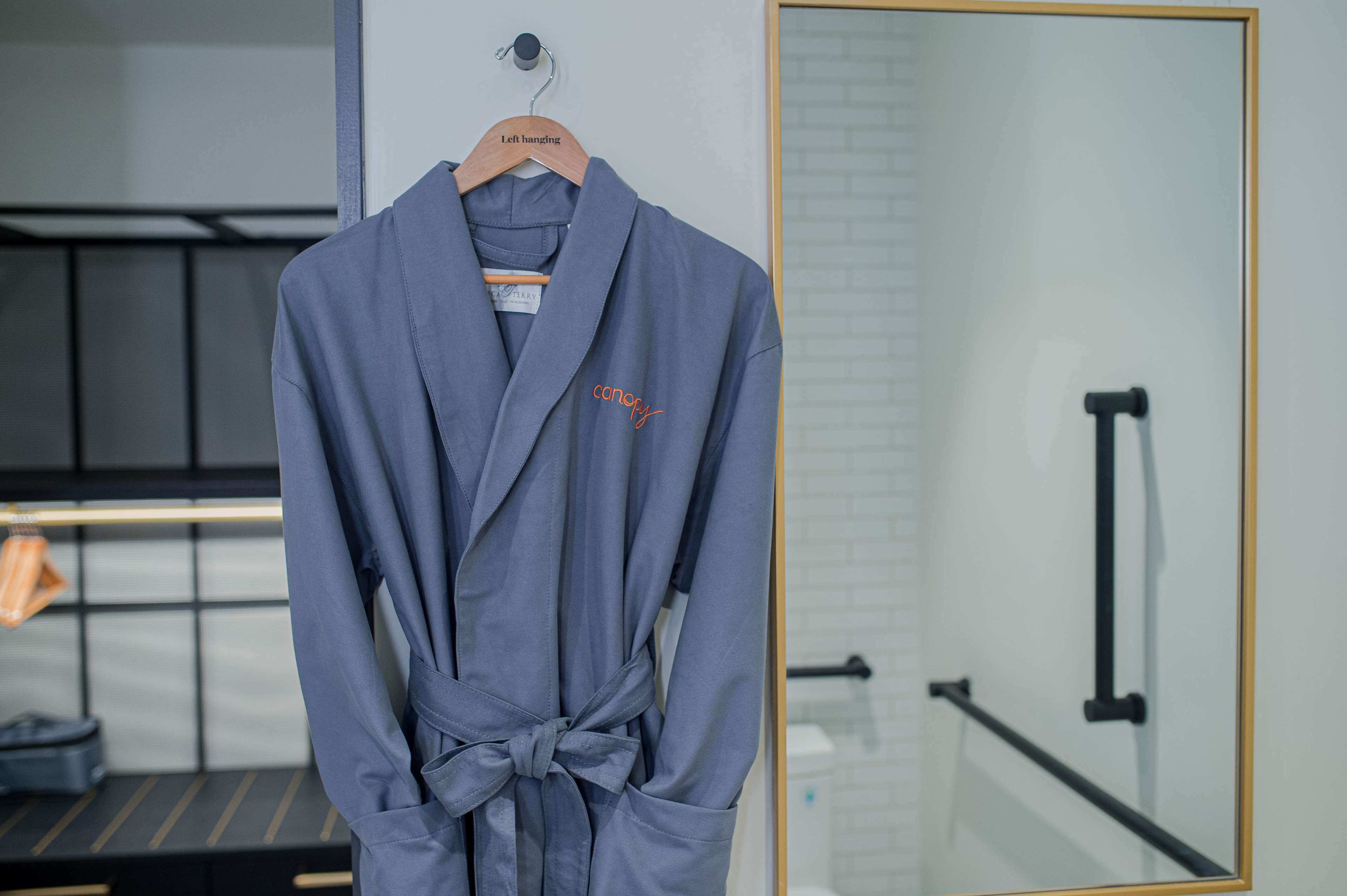 A grey robe hanging on a wooden hanger in a room with a bed, a black metal railing, and a white tiled wall.