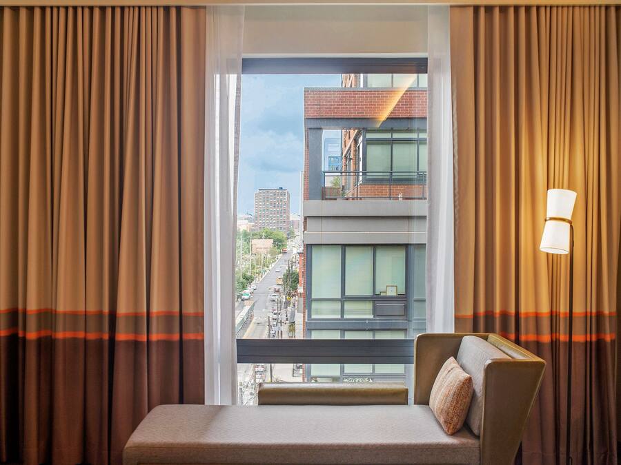 Habitación de hotel con vistas a un paisaje urbano a través de amplias ventanas, un sofá, una lámpara y cortinas con estampado a rayas.