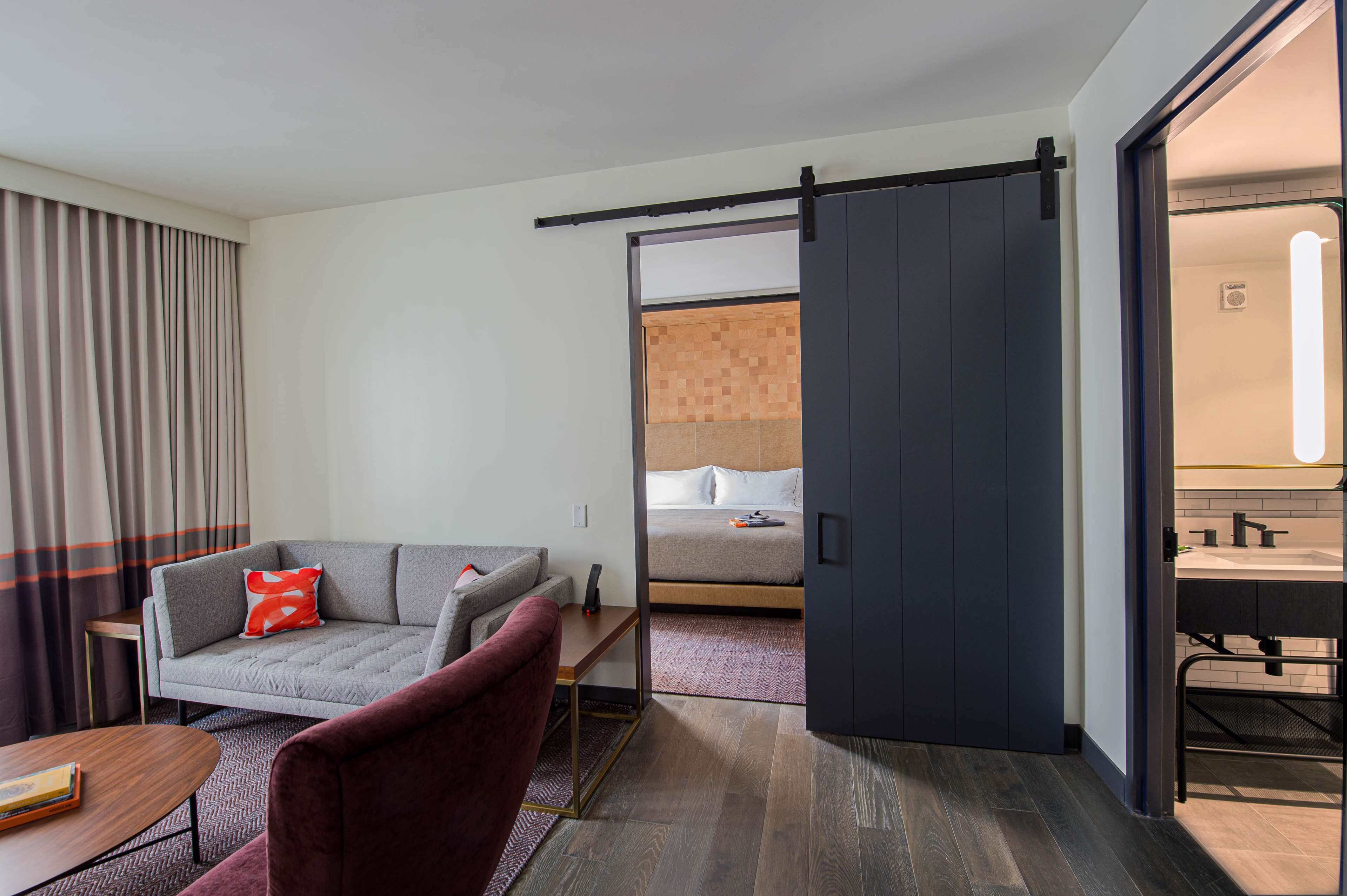 Ein Hotelzimmer mit einer Couch, einem roten Sessel, einem kleinen Tisch, einem Bett und einem Badezimmer mit Spiegel und Waschbecken.