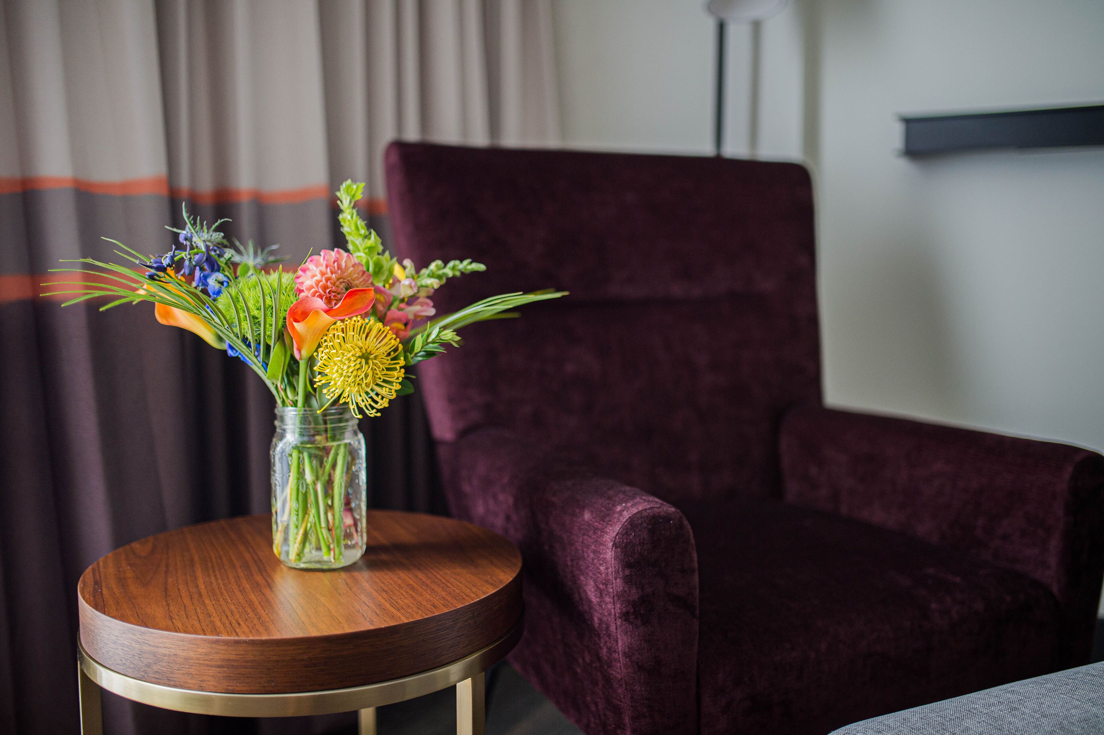 A room with a round wooden table holding a vase of flowers, a purple velvet chair, and a white wall with a black shelf.