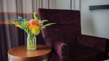 A room with a round wooden table holding a vase of flowers, a purple velvet chair, and a white wall with a black shelf.