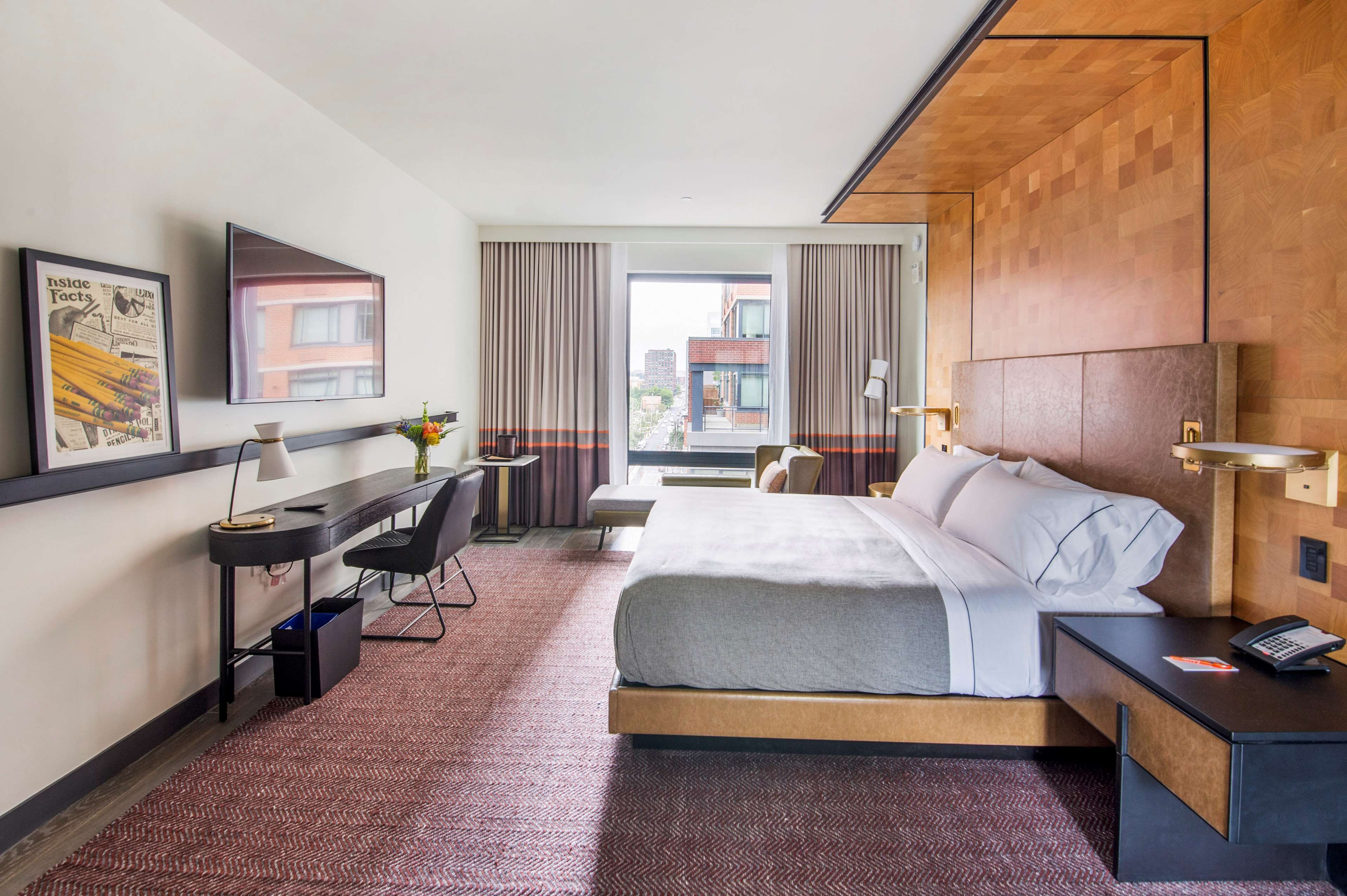 A hotel room with a large bed, a desk with a chair, a TV mounted on the wall, and a view of the city through the window.