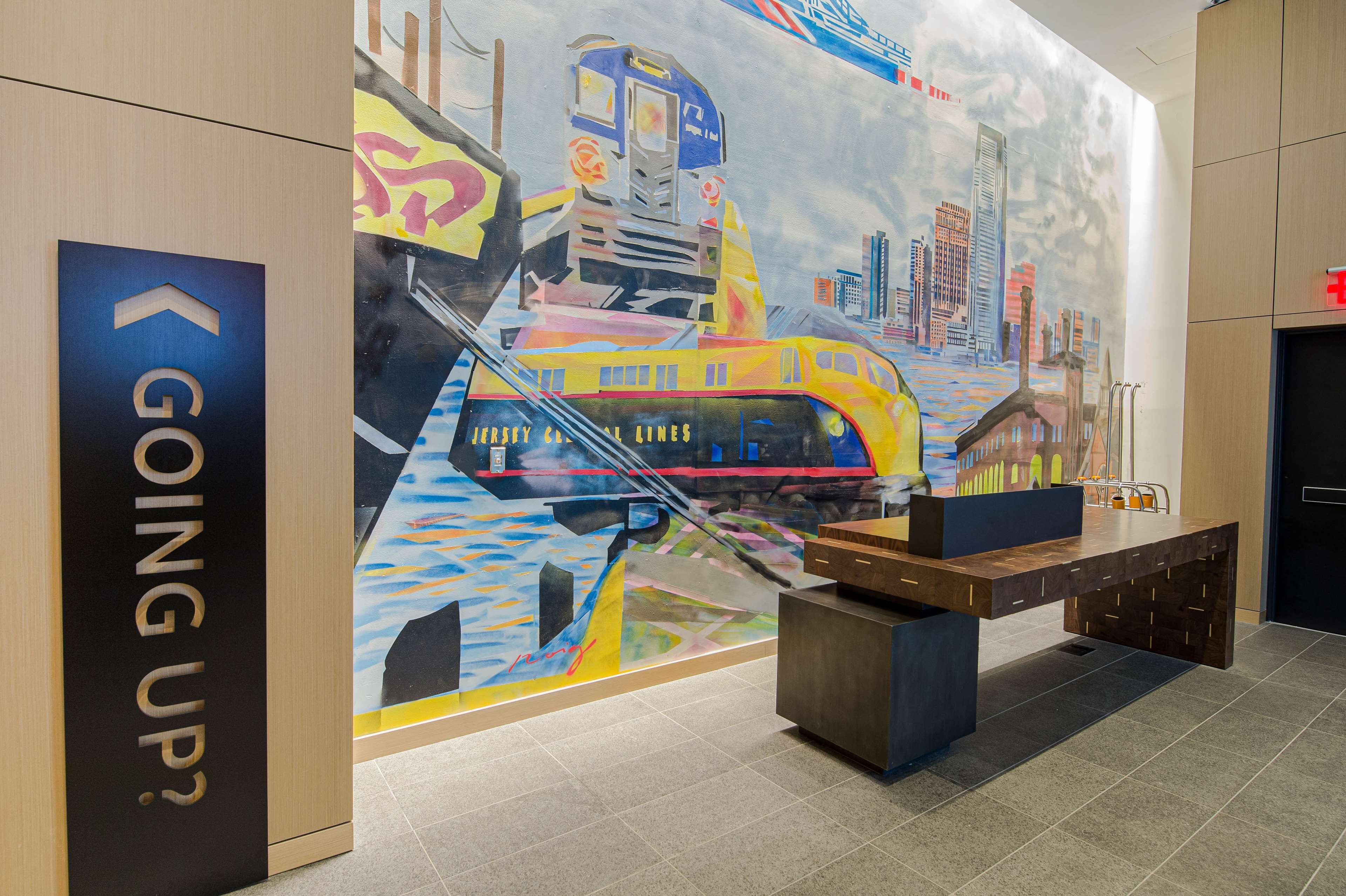 A modern lobby with a mural of a train, a reception desk, and a sign reading "GOING UP?".