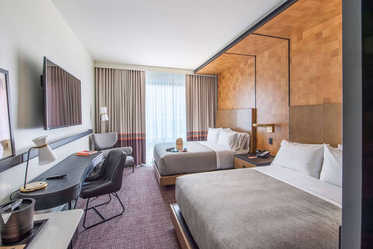 A hotel room with two beds, a desk with a chair, a TV, and a large window with curtains.