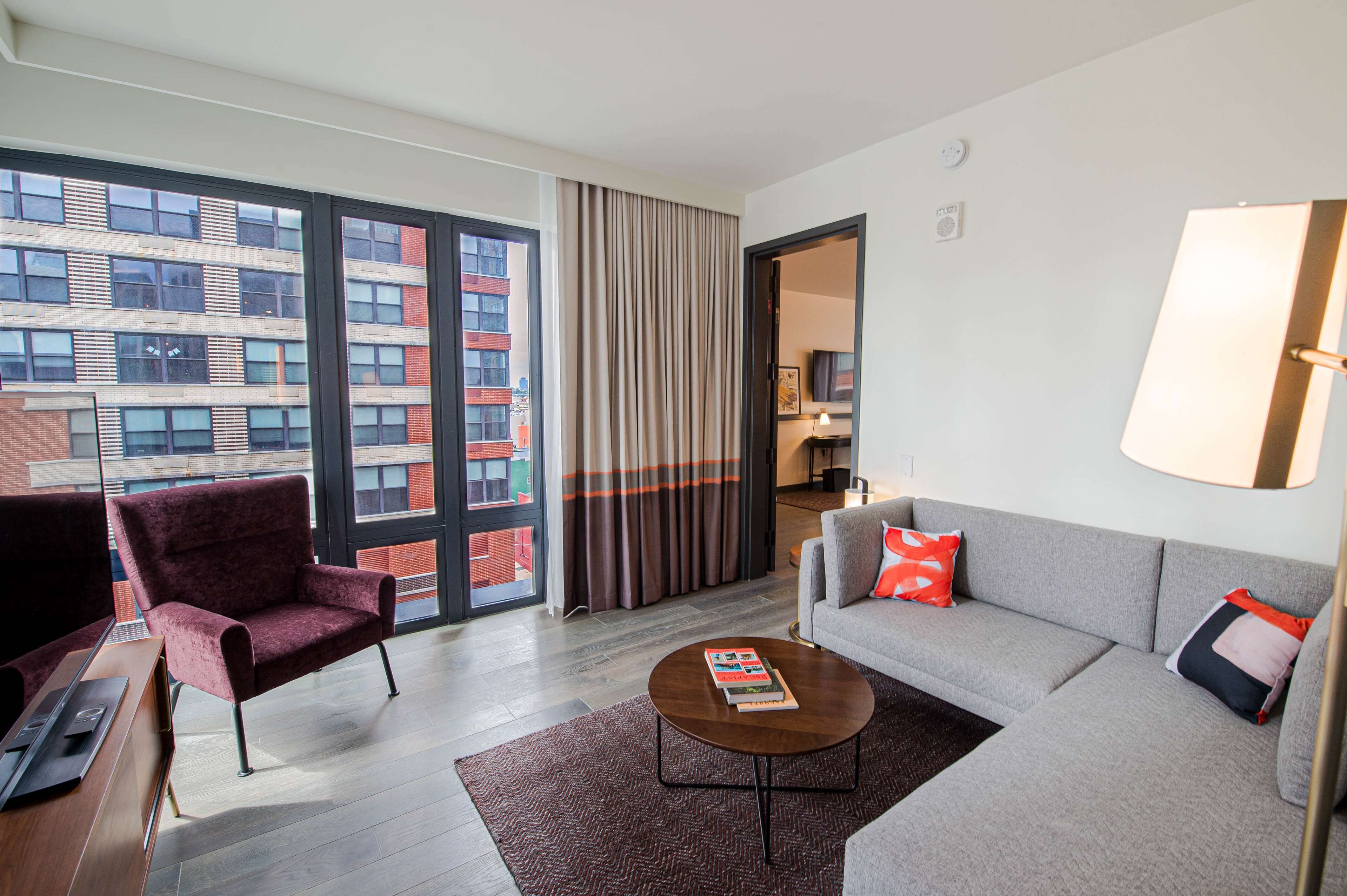 A modern hotel room with a large window, a grey sofa, a red armchair, and a coffee table.