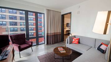 A modern hotel room with a large window, a grey sofa, a red armchair, and a coffee table.